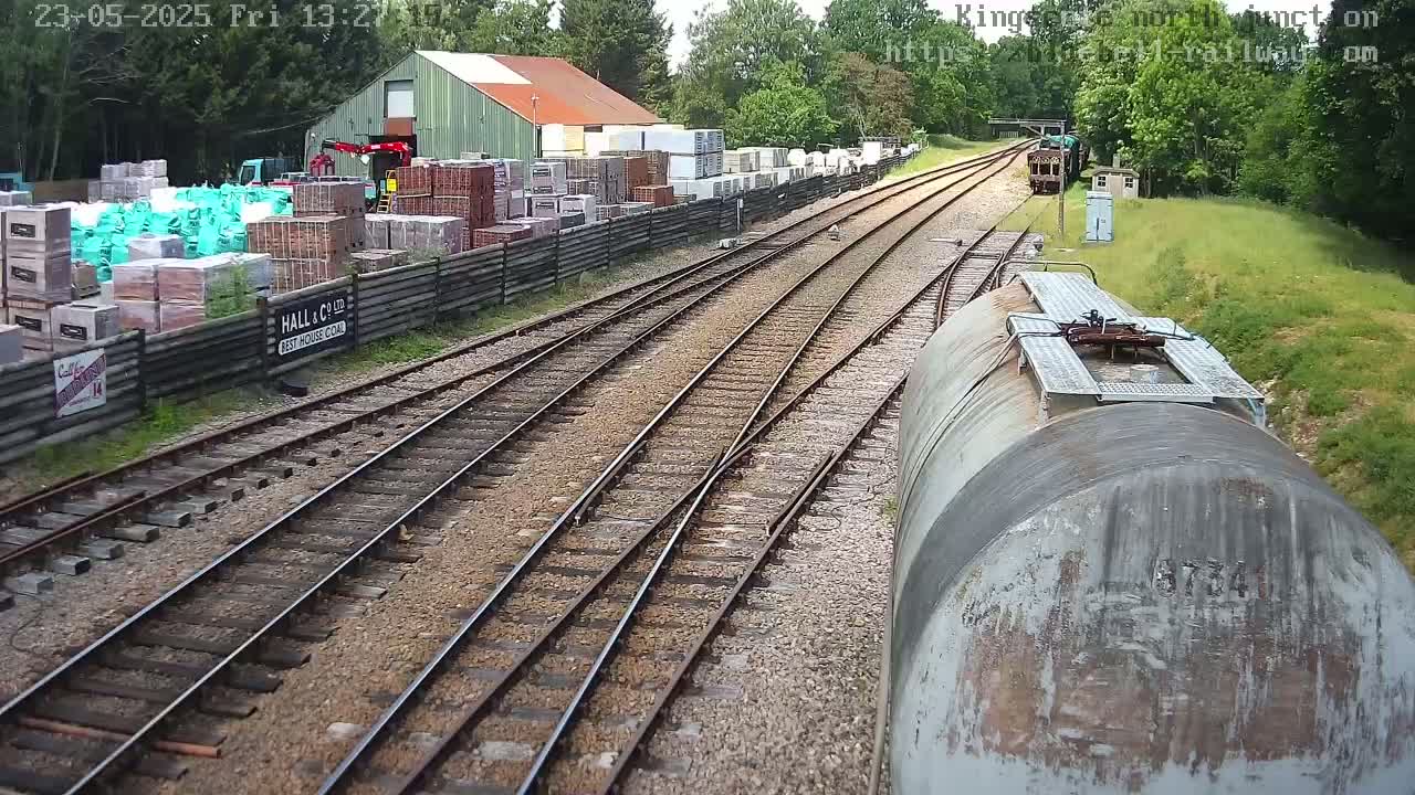 A view from a train car shows railway tracks branching out from a yard filled with stacks of building materials and a warehouse, under a partly sunny sky.