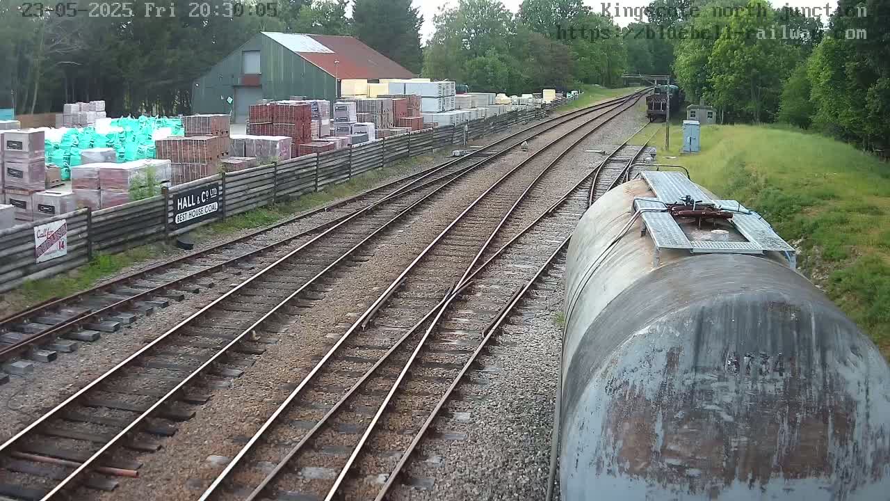 A view from a train shows several railway tracks converging near a building with stacked building materials beside a wooden fence, under a partly cloudy sky.