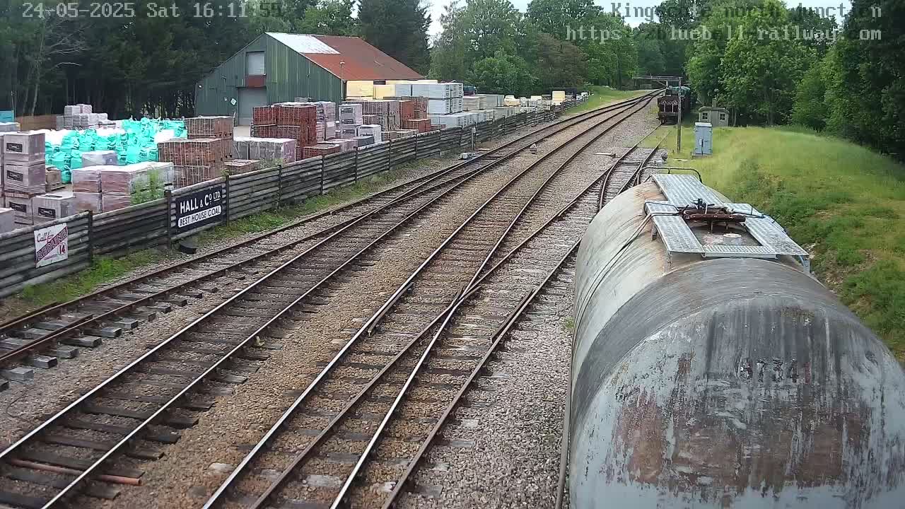 A view from a railway car shows a rail yard with multiple tracks diverging, a building containing stacks of building materials, and green vegetation under a partly sunny sky.