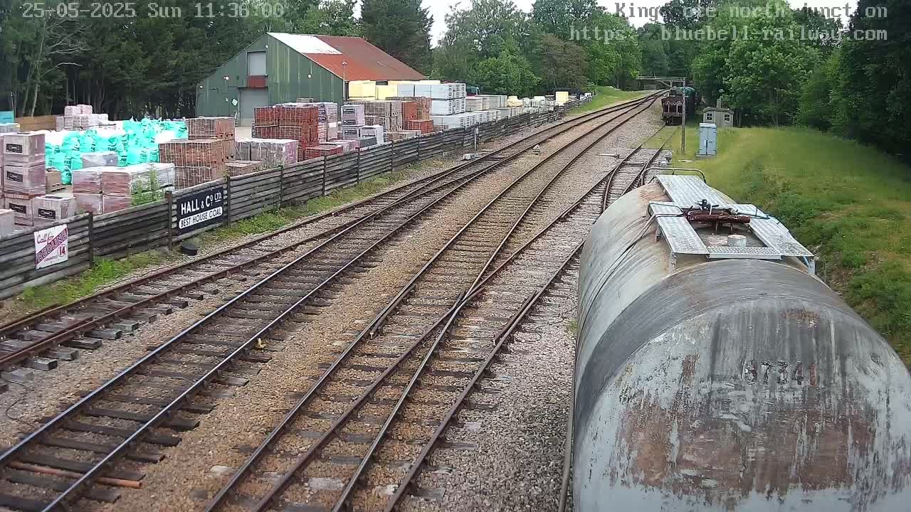 A railway junction on a sunny day with multiple tracks, a weathered tanker car, and a building stocked with bricks and other building materials in the background.