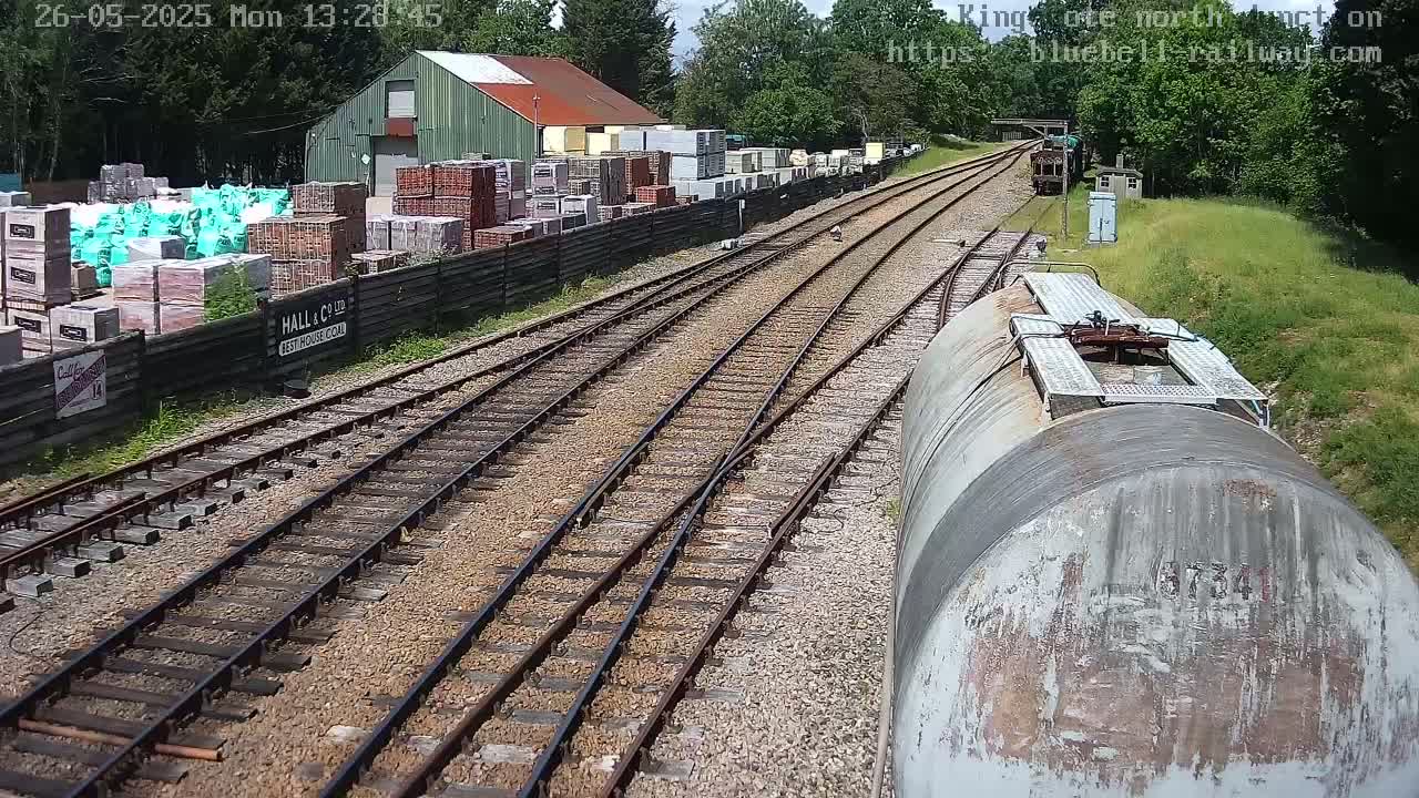 A view from a train of railway tracks splitting into two directions, passing a building with stacked bricks and bags, under a sunny sky.