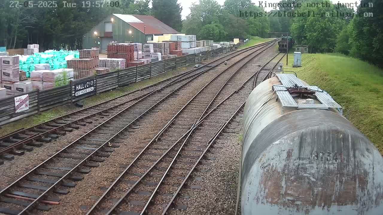 A railway junction with several tracks, a building storing stacks of bricks and other building materials, and a large metal tank car are visible, all under a cloudy sky.