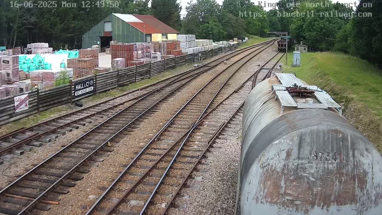 A view from a railway car shows a railway junction with multiple tracks, a building storing building materials, and some trees in the background on a partly cloudy day.