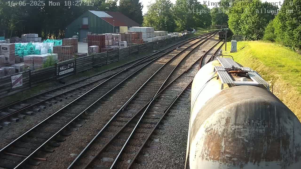 A train travels past a building materials yard on a sunny day, alongside railway tracks that split into multiple directions.