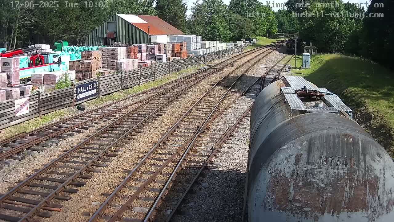 A weathered tanker car sits on a railway track on a sunny day, overlooking a yard filled with stacks of building materials and a red barn.