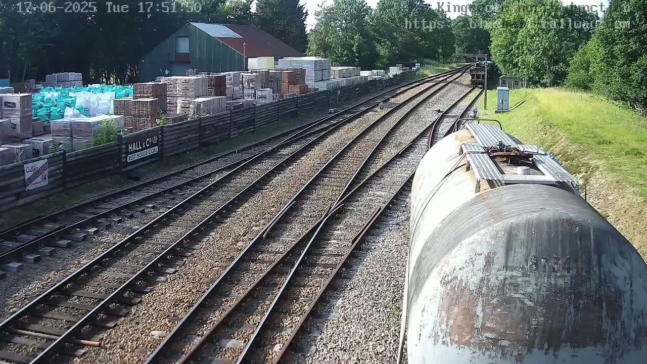 A view from a train shows multiple train tracks branching off, alongside a building and a yard stacked with building materials, all under a sunny sky.
