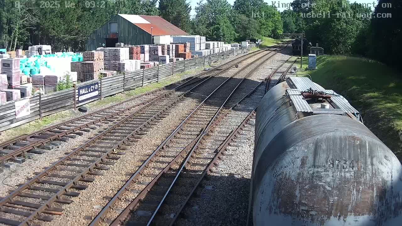 A view from a train shows railroad tracks splitting into multiple paths, alongside a building with stacked building materials, all under a sunny sky.