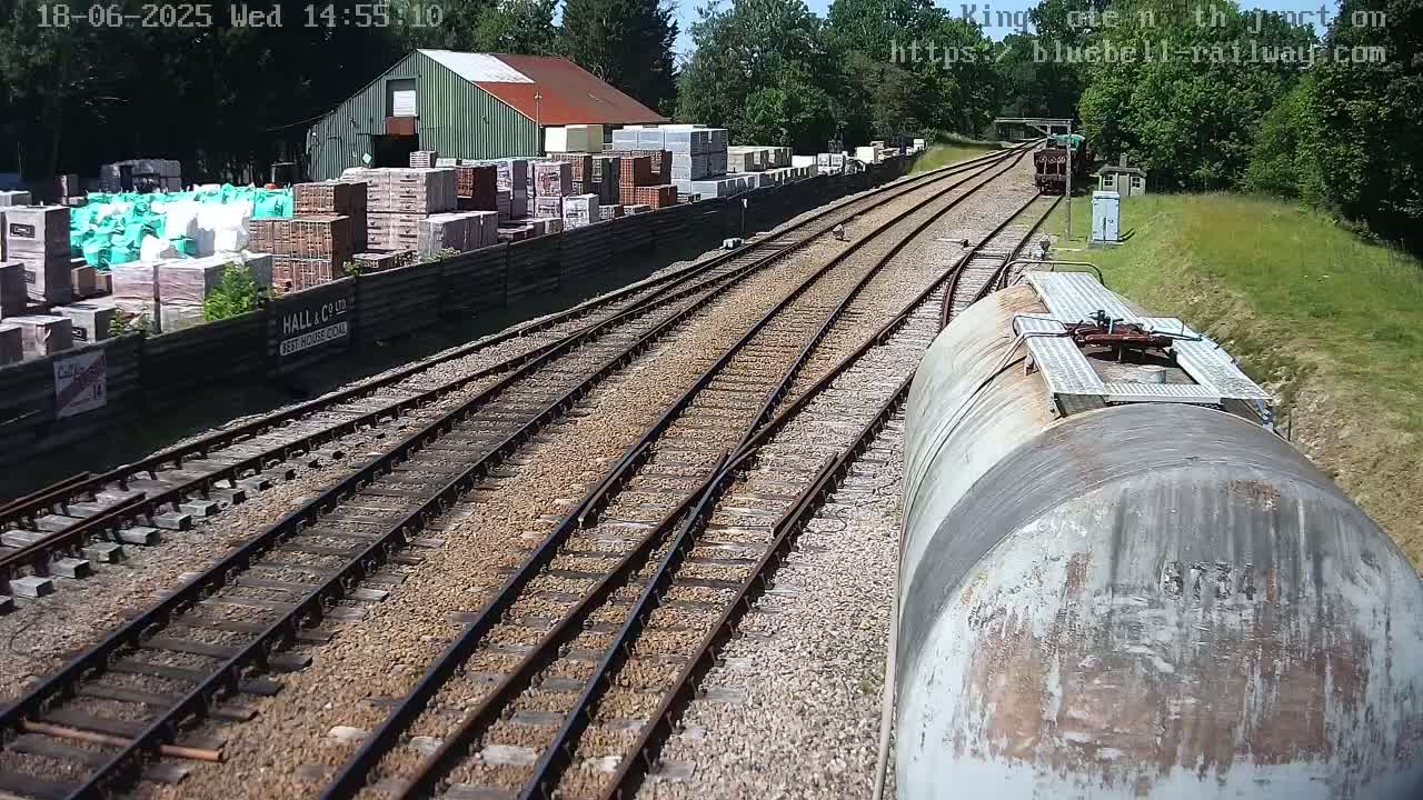 A view from a train shows railway tracks splitting into multiple lines, passing alongside a building and stacks of building materials, under a sunny sky.