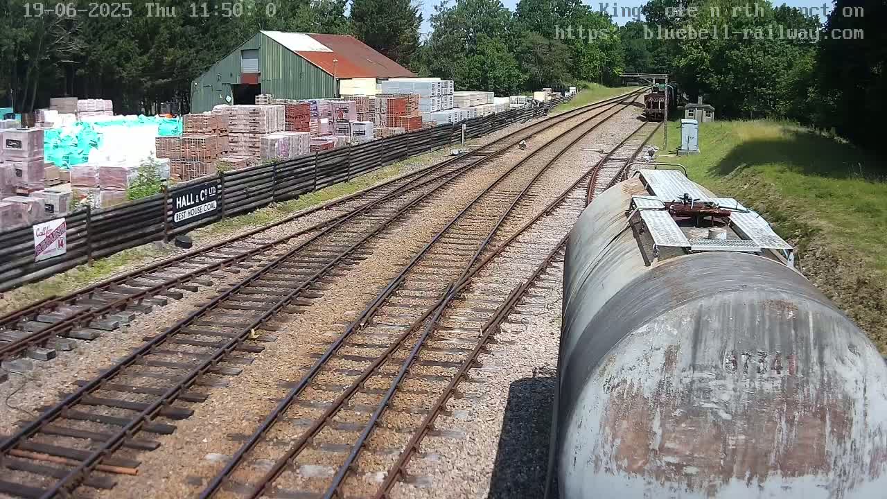 A view from a train car shows railroad tracks splitting into two, passing alongside a building and a stack of bricks in a sunny, outdoor setting.