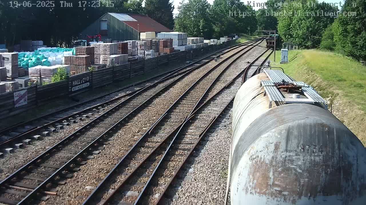 A view from a train car shows railway tracks splitting into multiple directions, alongside a building with stacked building materials and trees, on a sunny day.