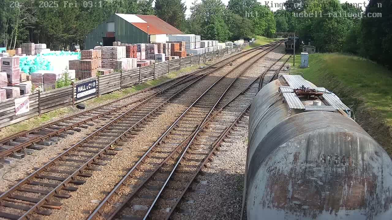 A view from a train shows railway tracks splitting, with a building and stacks of building materials to the left, and a grassy embankment to the right, on a sunny day.
