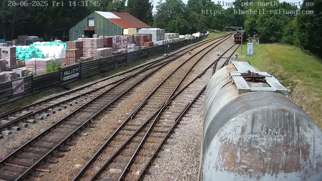 A view from a train shows a railway line splitting into two, with a weathered storage tank in the foreground, a building and stacks of materials beyond a fence on the left, and trees lining the right side on a sunny day.