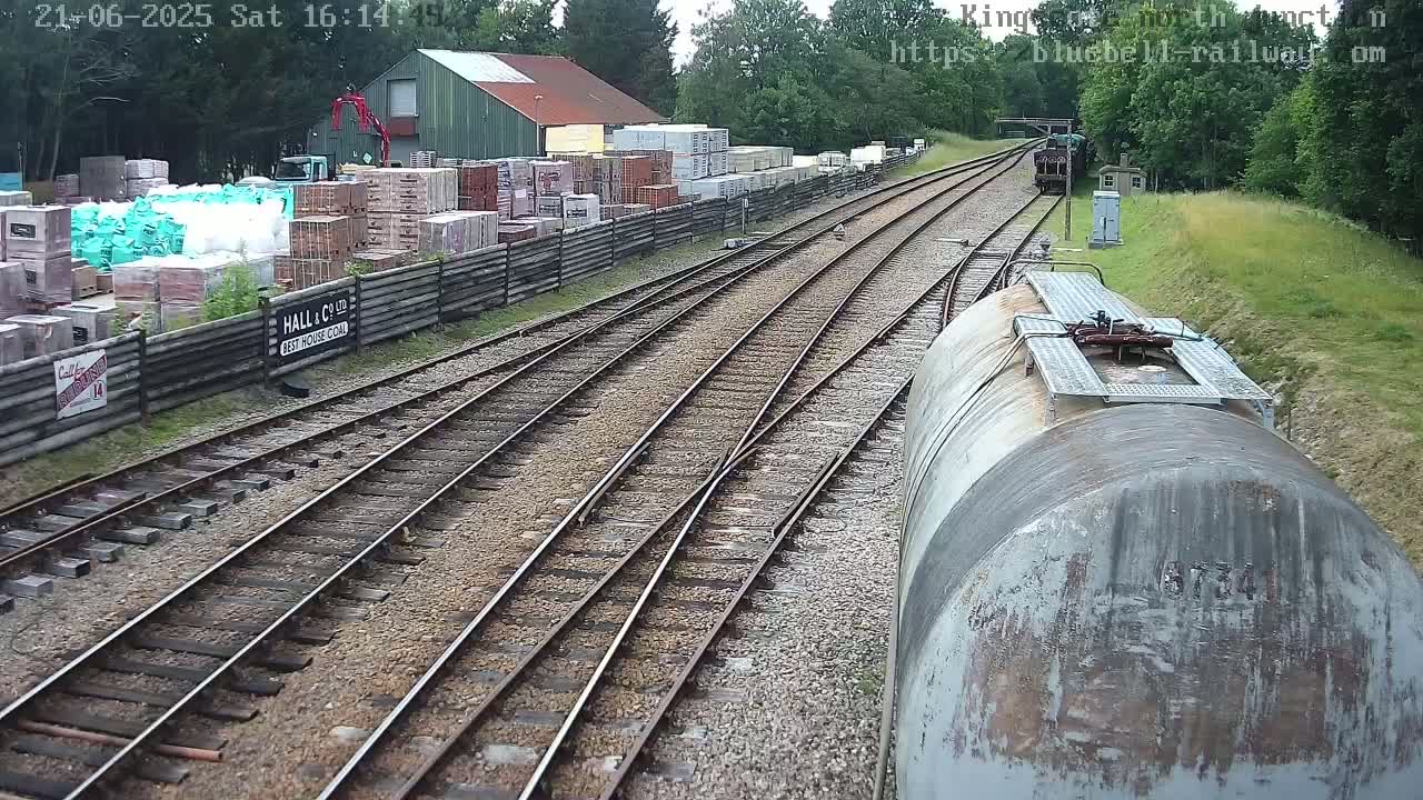 A railway line with multiple tracks curves through a yard with stacked building materials, under a partly sunny sky.