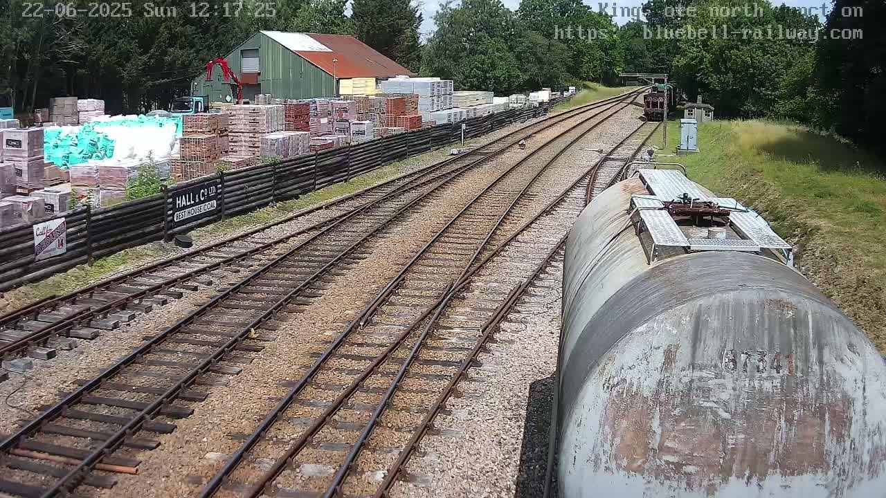 A view from a railway car shows train tracks splitting into two directions, passing by a building materials yard on a sunny day.