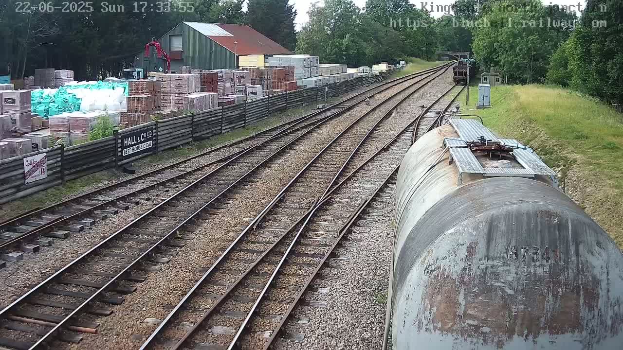 Railway tracks diverge near a building materials yard on a sunny day, with a train car visible in the foreground.