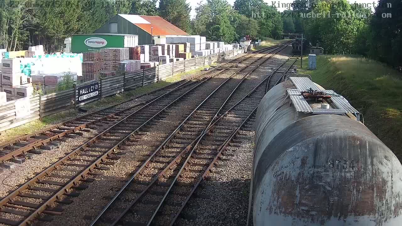 A train car sits on a railway track next to a yard with stacked building materials, under a sunny sky.