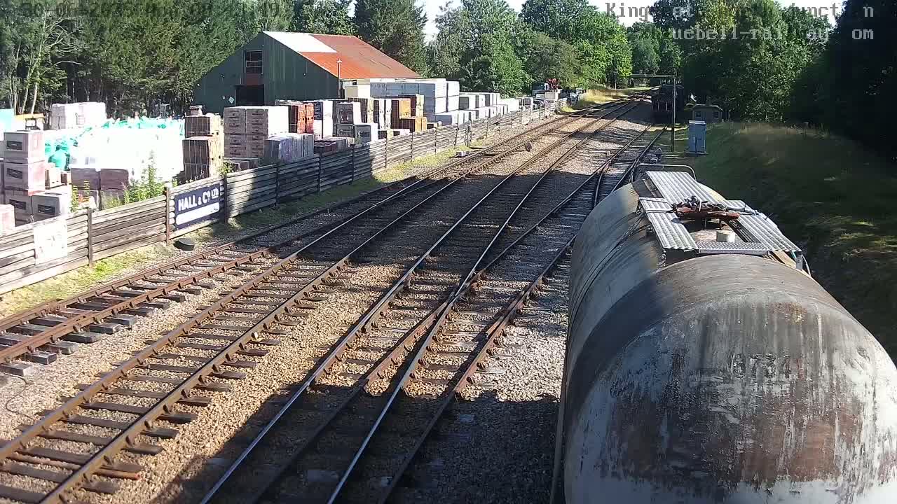 A view from a train car shows multiple train tracks splitting and converging, alongside a building and stacks of building materials, all under a sunny sky.