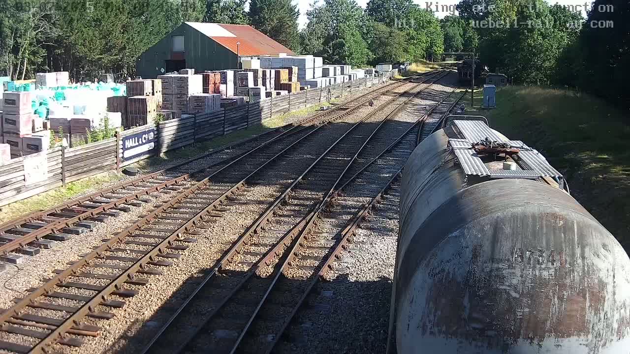 A train's view of railway tracks branching out from a yard on a sunny day, with building materials stacked along a fence beside the tracks.
