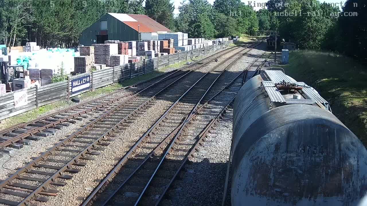 A view from a train shows railway tracks splitting, passing by a building and a yard full of stacked building materials, on a sunny day.