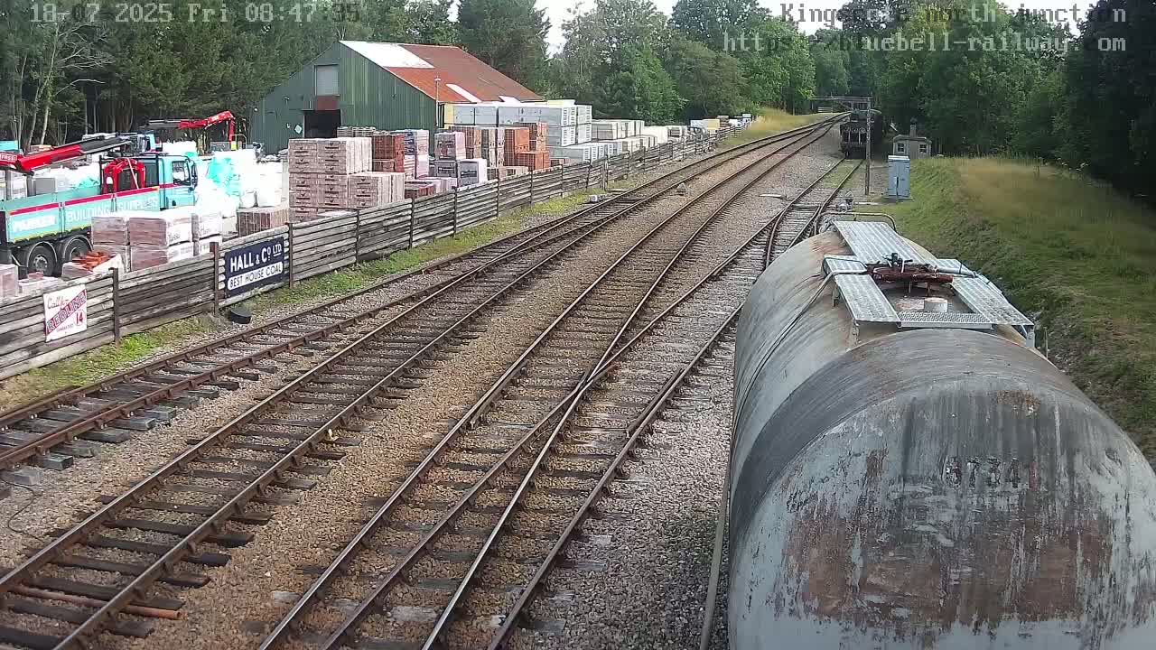 A train car overlooks railway tracks that diverge near a building and a yard with stacked building materials, under a partly sunny sky.