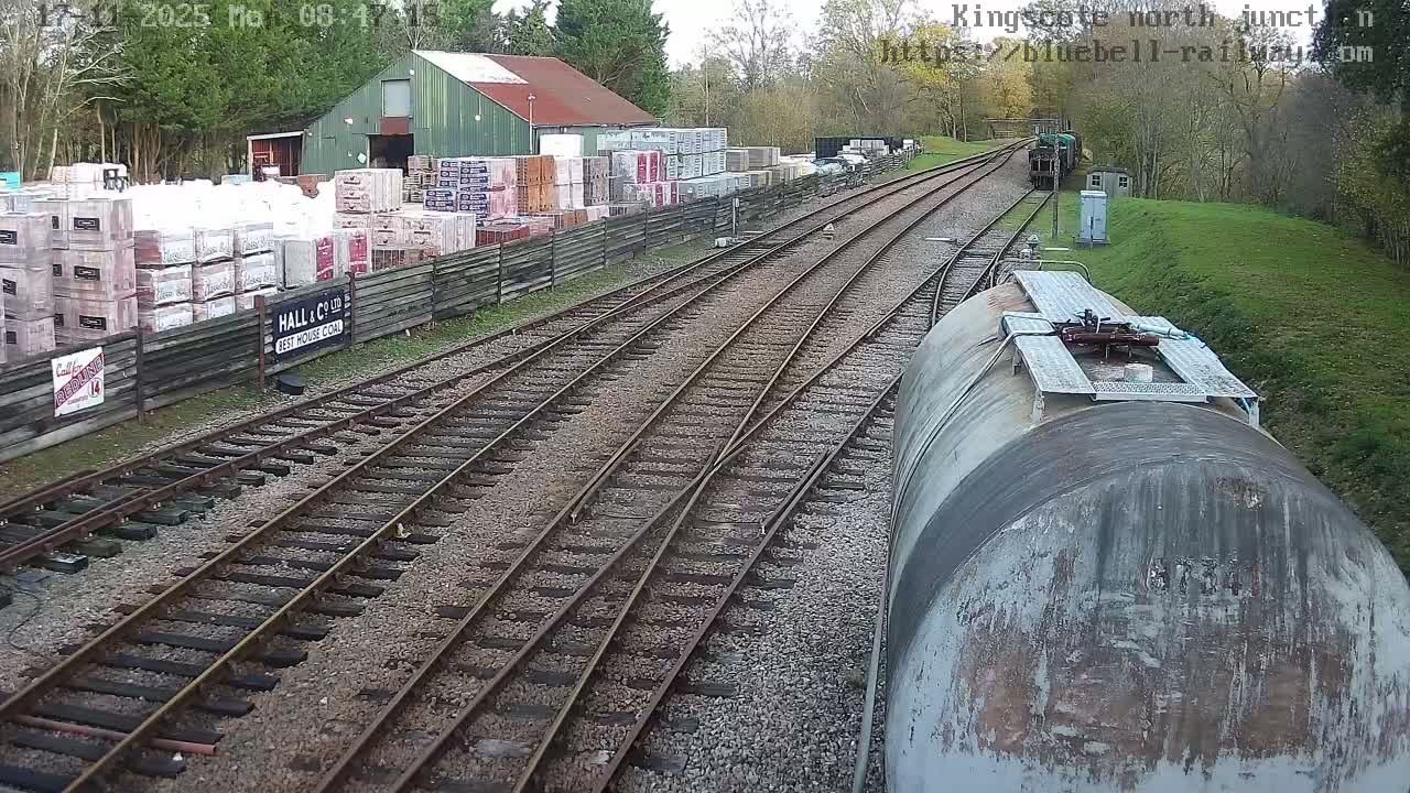 On an overcast day, a high-angle view captures multiple railway tracks curving into the distance, with a weathered tank car in the foreground, an industrial yard piled with various materials next to a green-roofed building on the left, and a green locomotive visible further down the tracks.