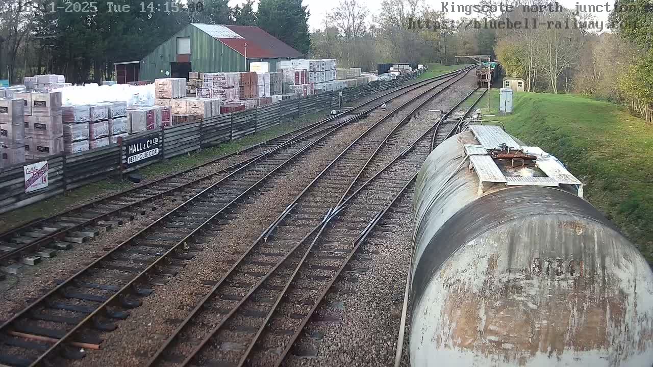 On an overcast day, a high-angle view captures multiple railway tracks curving into the distance, with a weathered tank car in the foreground, an industrial yard piled with various materials next to a green-roofed building on the left, and a green locomotive visible further down the tracks.