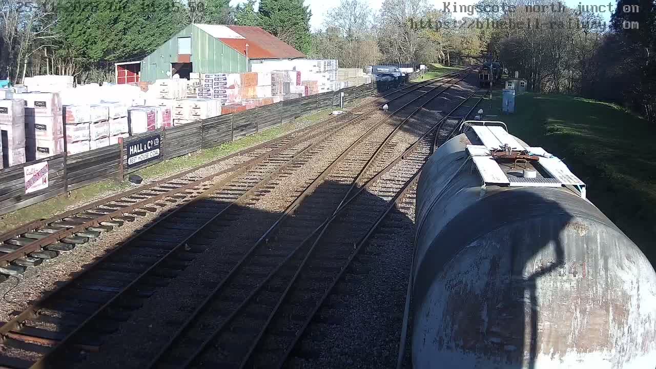 On a sunny day, multiple railway tracks, including a large grey tank car on the right, extend towards a distant train, with industrial buildings and stacked goods lining the left side.