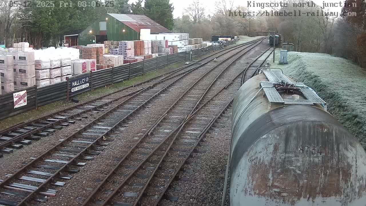 A frosty railway yard features multiple tracks curving into the distance, a storage area stacked with materials beside a green-roofed building on the left, and the weathered top of a large train car visible in the foreground on the right.