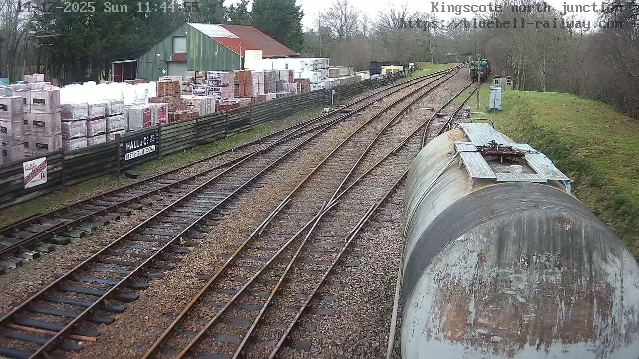 A frosty railway yard features multiple tracks curving into the distance, a storage area stacked with materials beside a green-roofed building on the left, and the weathered top of a large train car visible in the foreground on the right.