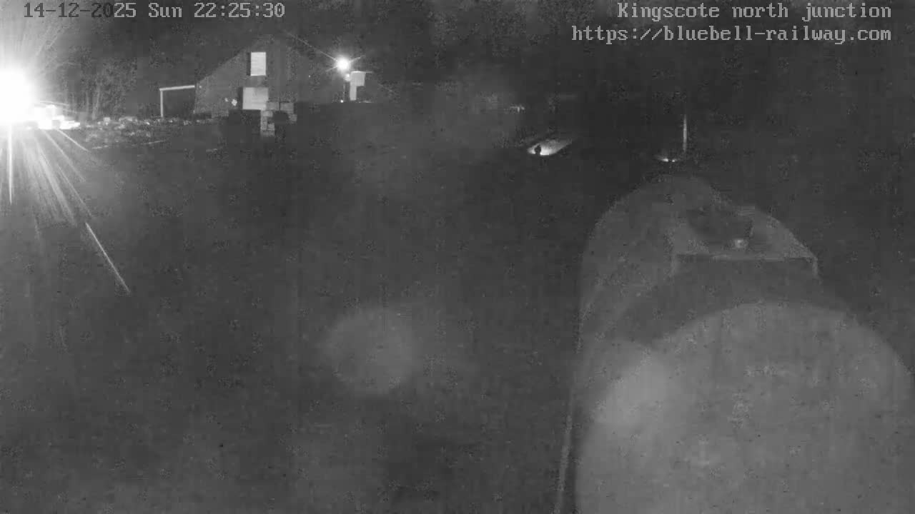 A frosty railway yard features multiple tracks curving into the distance, a storage area stacked with materials beside a green-roofed building on the left, and the weathered top of a large train car visible in the foreground on the right.