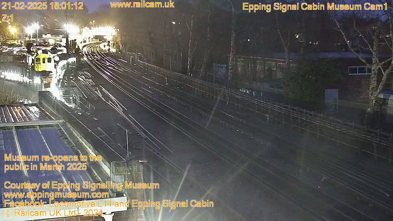 A nighttime view of a train station with a yellow train visible and several sets of train tracks, under wet conditions.