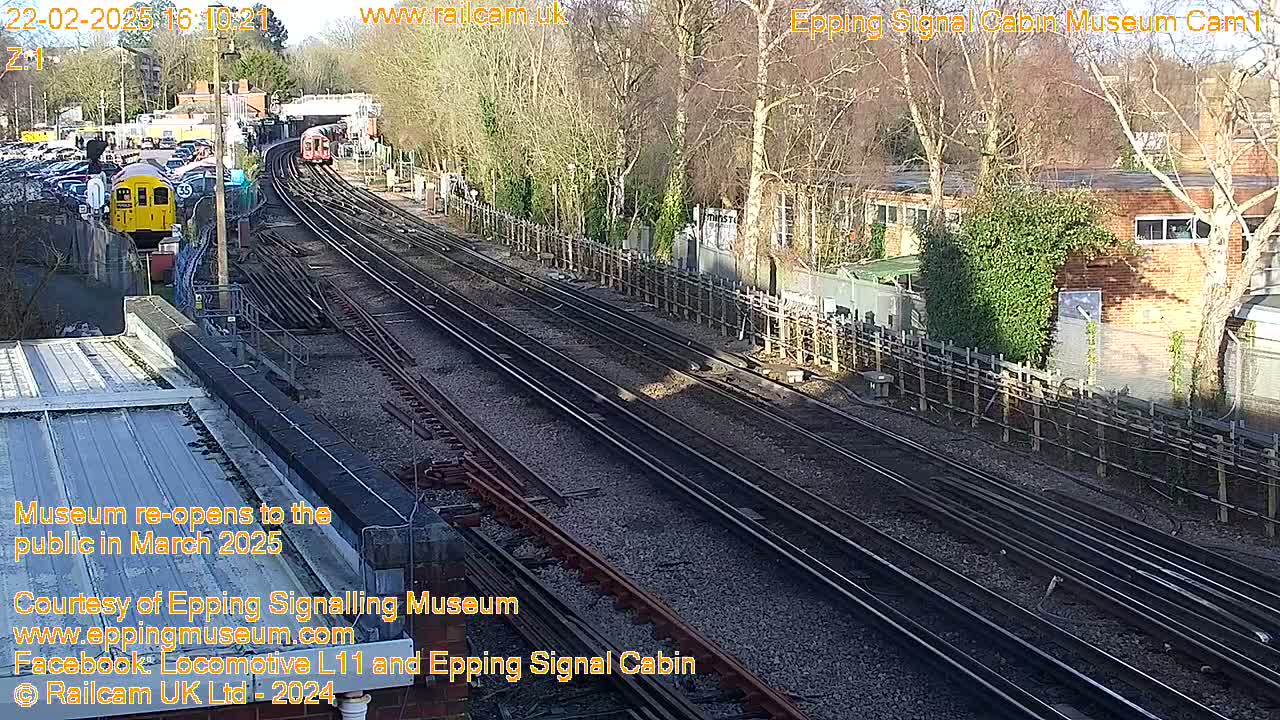 Two trains are visible on multiple railway tracks next to a building, under a sunny sky.