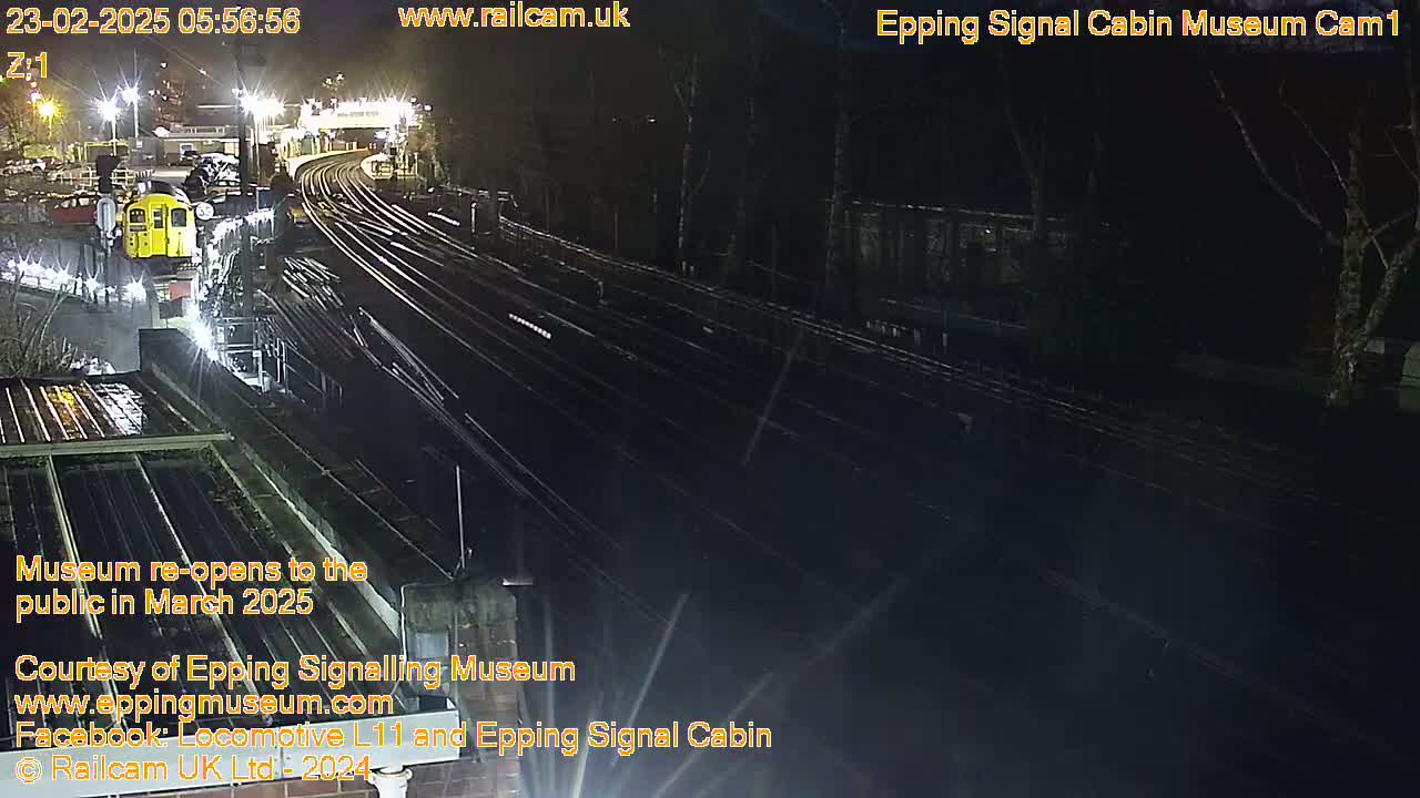A nighttime view of a train station with a train and railway tracks, under wet conditions.