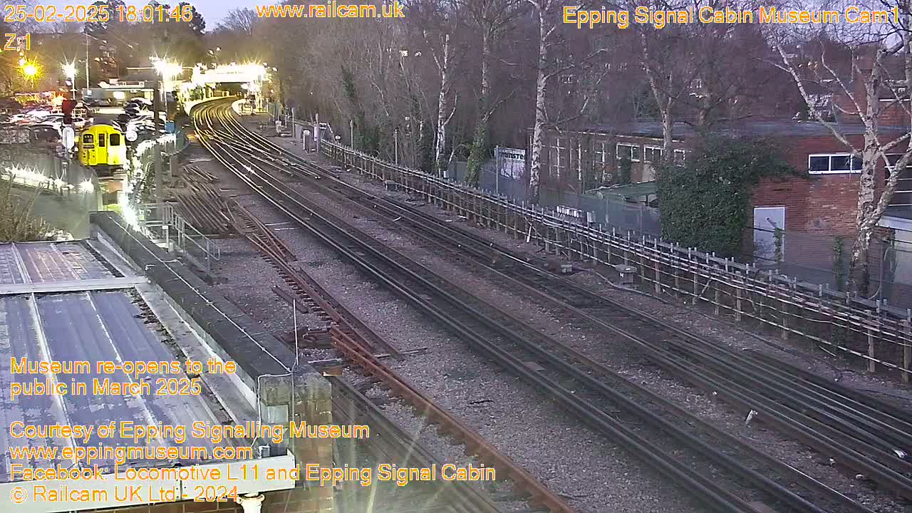 A twilight view of a train station with multiple train tracks, a yellow train car, and bare trees surrounding a brick building.