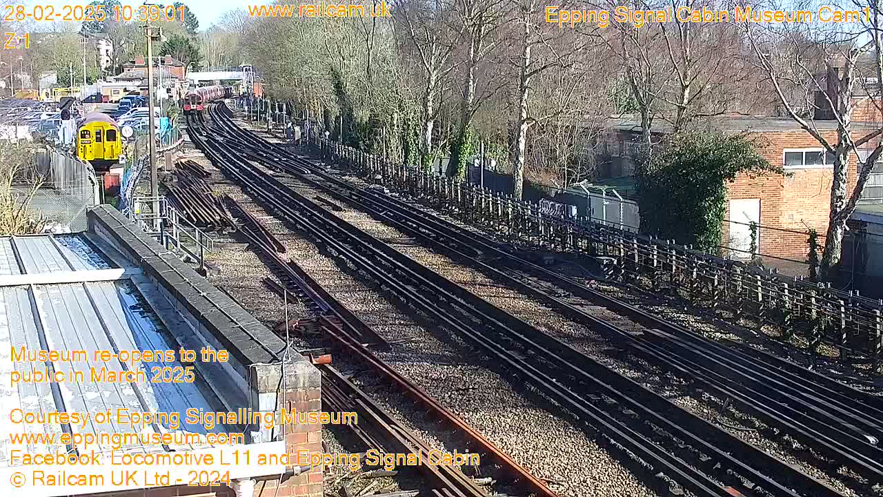 A yellow train is visible in the distance on a sunny day, with multiple train tracks stretching into the background, surrounded by bare trees and a brick building.