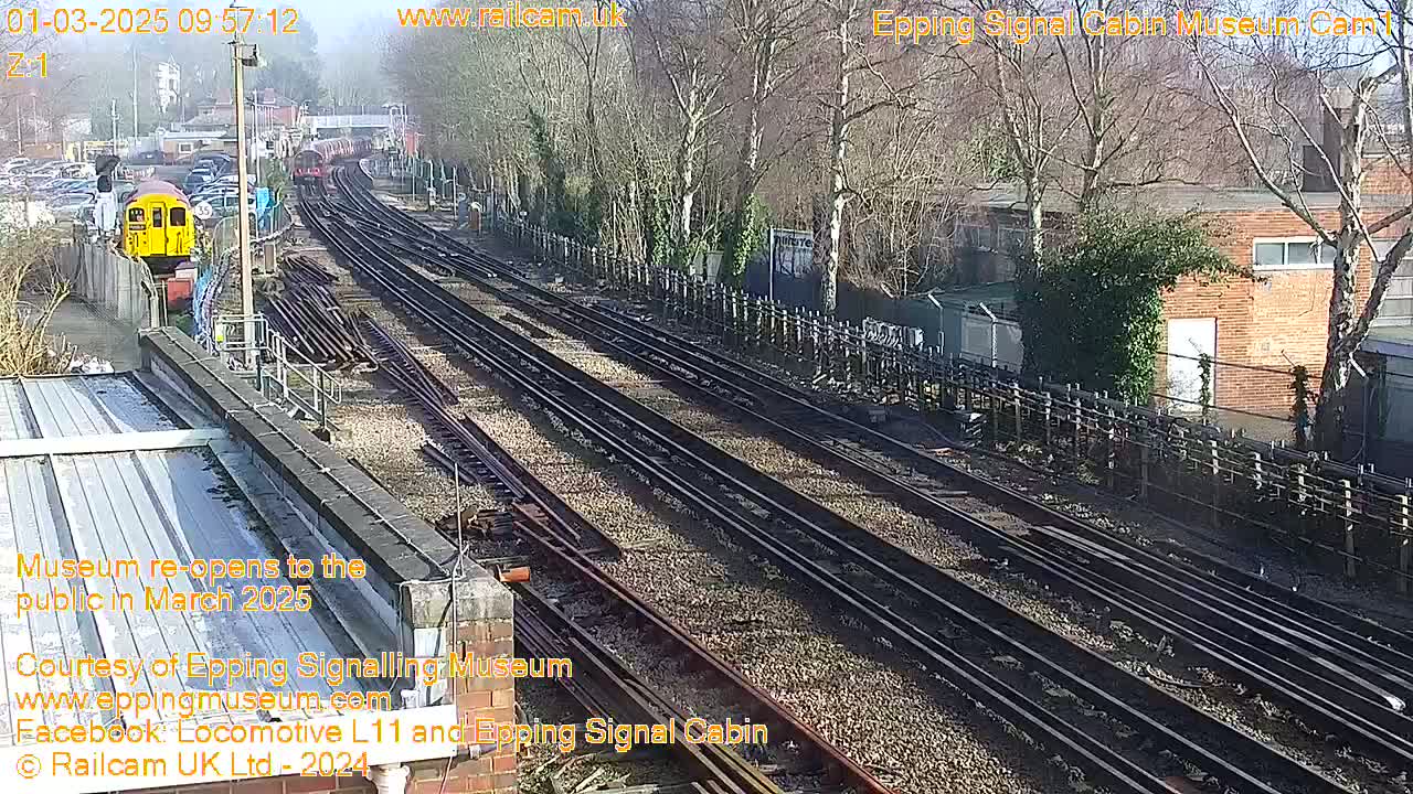 A sunny day at a railway yard shows multiple train tracks, a yellow train car, and buildings in the background.