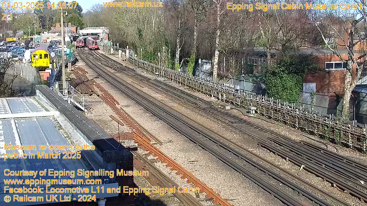 A sunny day shows multiple train tracks with two vintage train cars visible, near a brick building and wooded area.