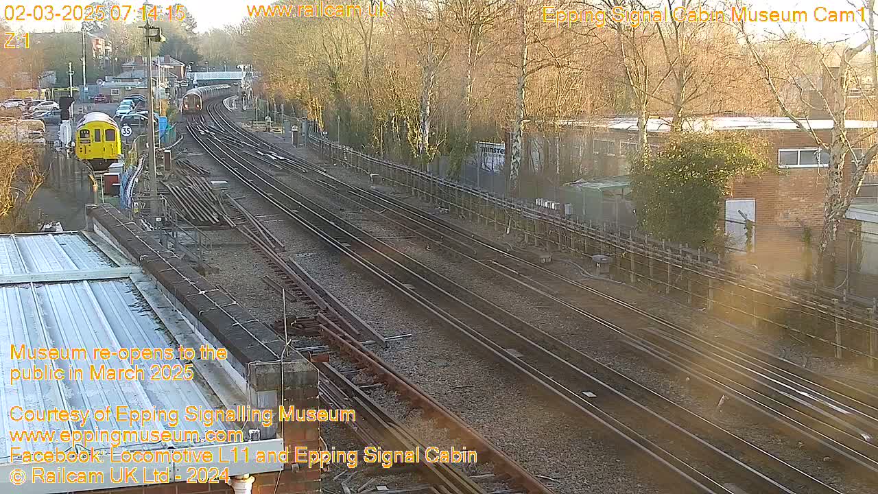 A yellow train passes by multiple train tracks on a sunny day, near a brick building and wooded area.