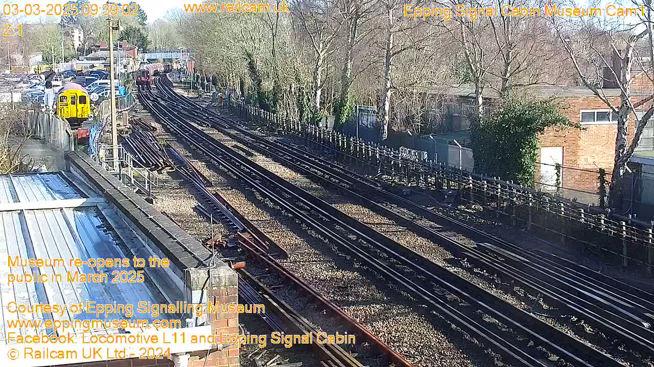 A sunny day reveals a high-angle view of multiple train tracks, a yellow train car, and surrounding buildings and trees.