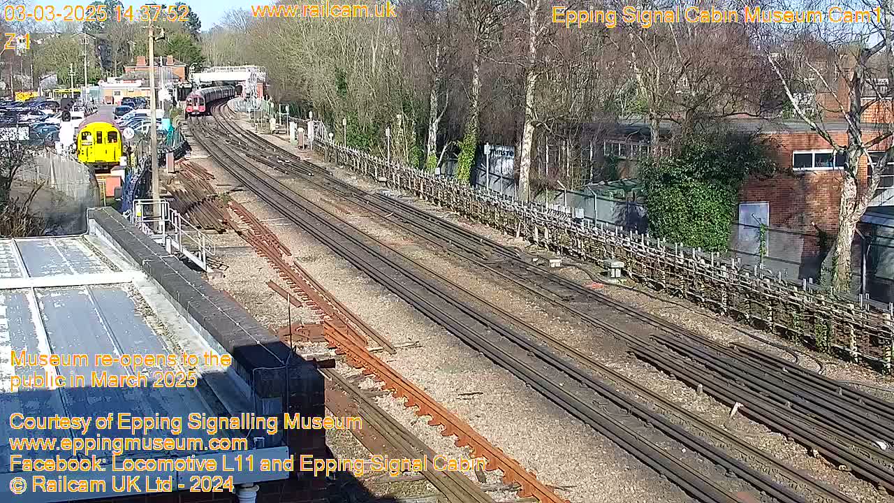 A sunny day at a railway station shows multiple train tracks, a yellow train, and a building.