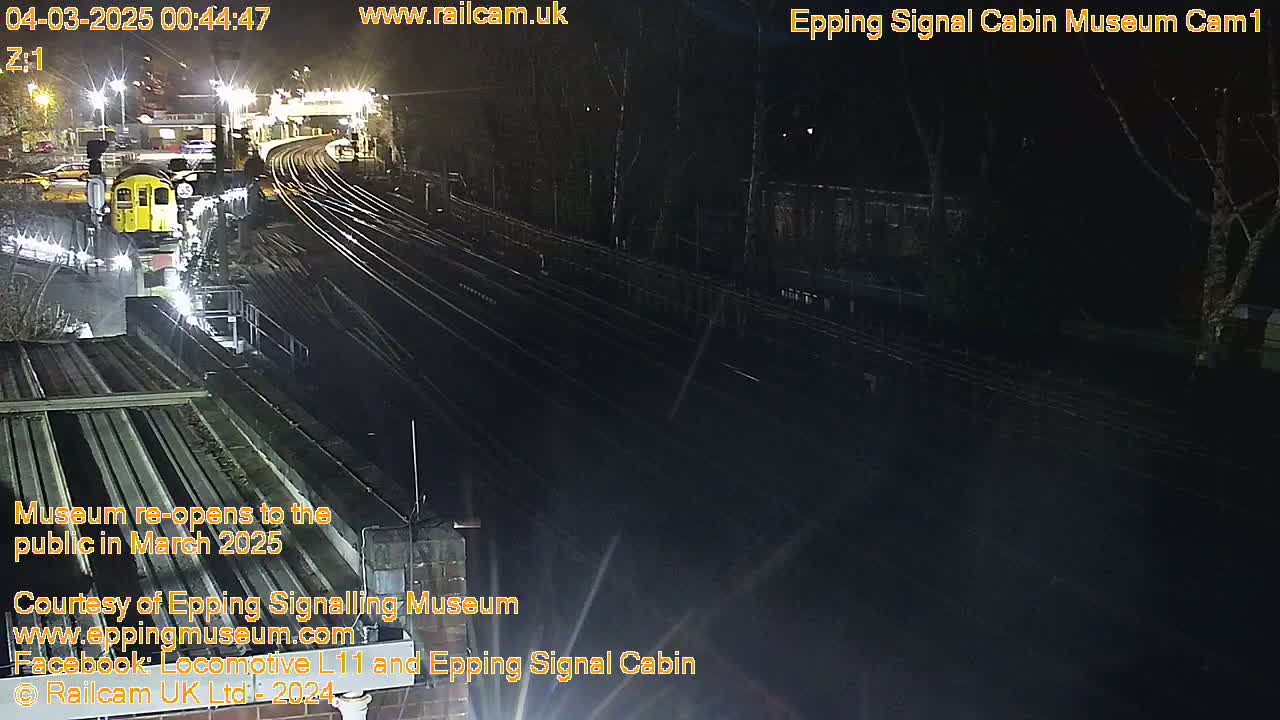 A nighttime view of a train station with a single train car visible, several railway tracks, and surrounding trees under dark conditions.