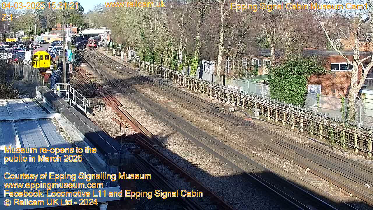 A sunny day at a railway station shows several train tracks, a yellow train car, and a brick building beyond a wooden fence.