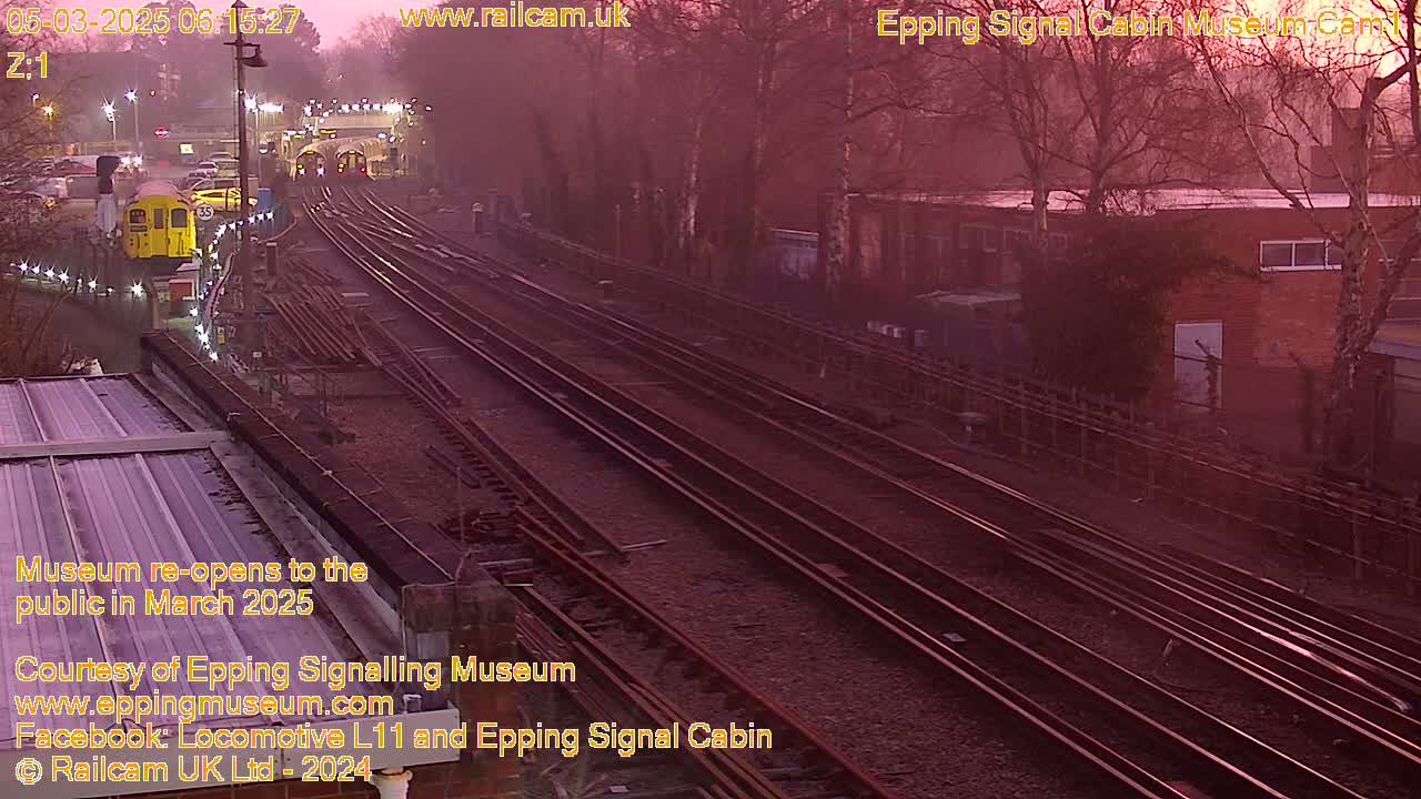 A hazy sunrise illuminates a railway station with multiple train tracks, a lone train car, and bare trees in the background.