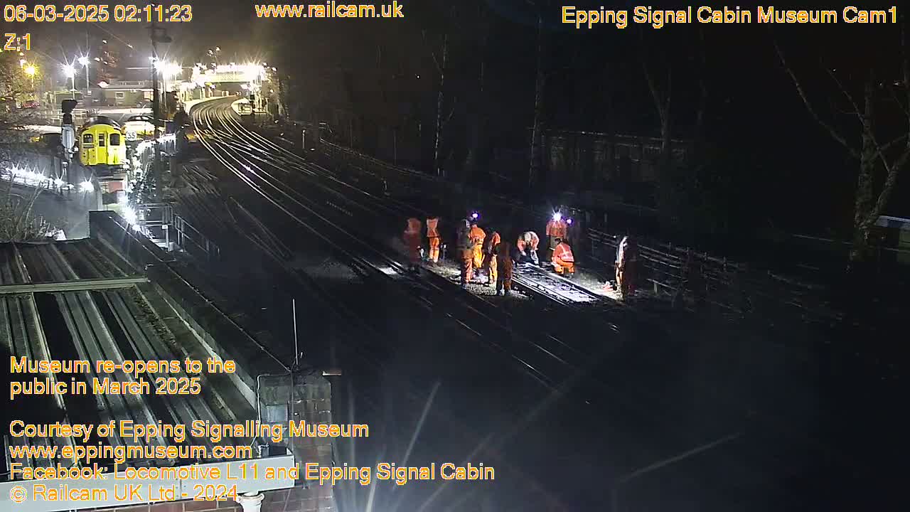 At night, several workers in orange are on the railroad tracks near a train and under bright lights.