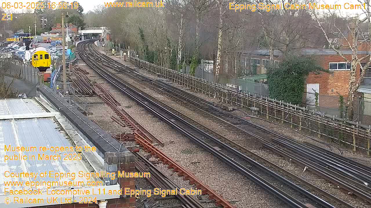 A yellow train is parked near a railway station on a mostly clear day, with multiple train tracks extending into the distance past a brick building and a line of trees.