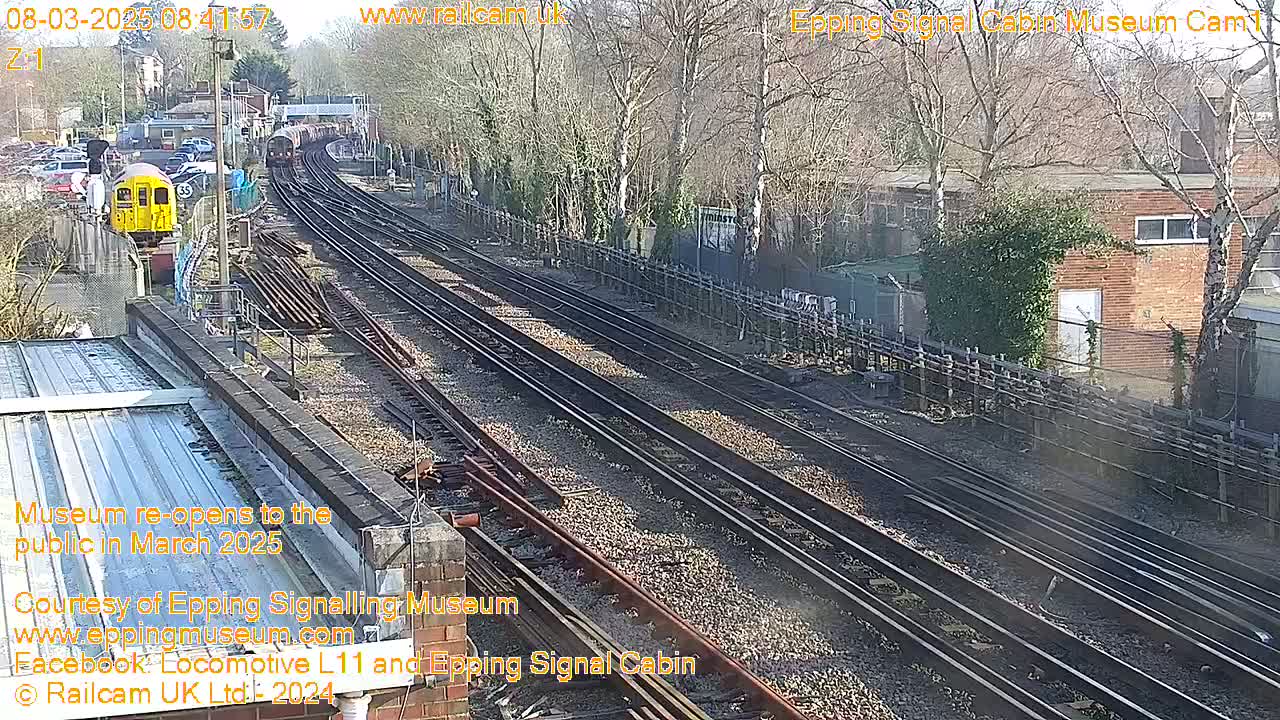 A sunny day shows multiple train tracks with a train and a yellow railway car visible, alongside buildings and trees.