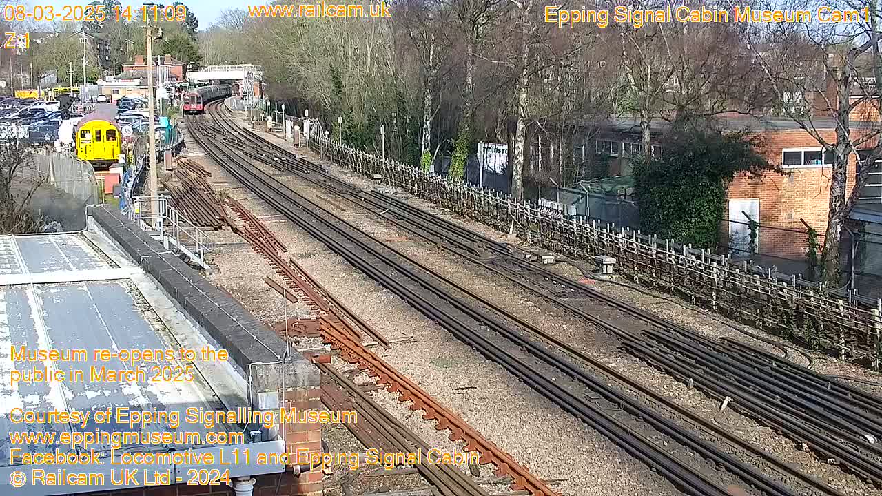 A sunny day at a railway station shows multiple train tracks, a yellow train car, and a brick building beyond a wooden fence.