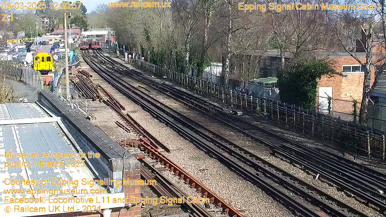 A sunny day reveals a railway yard with multiple train tracks, several parked trains, and surrounding buildings.