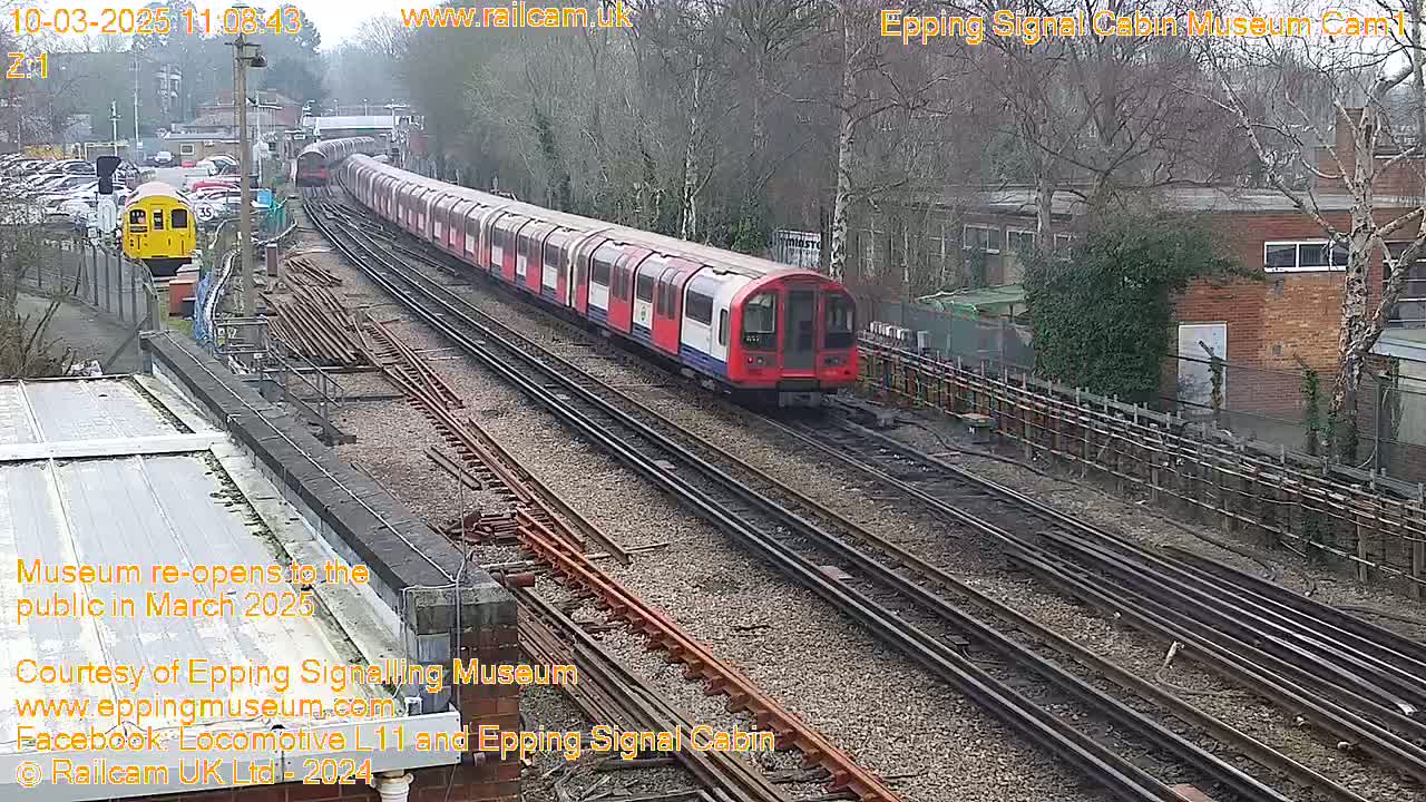 A long red and white train travels along multiple railway tracks on an overcast day, passing a smaller yellow train and a brick building.