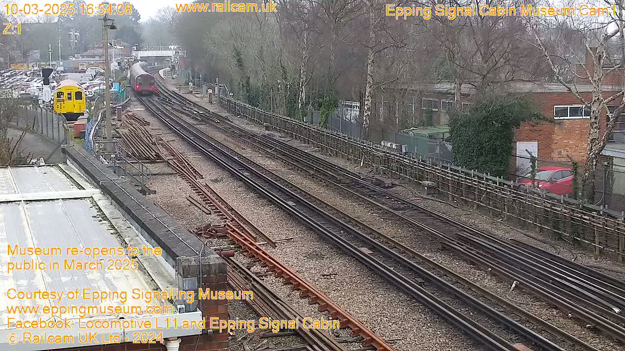 A train passes by a railway station on an overcast day, with multiple train tracks visible and a yellow train parked nearby.